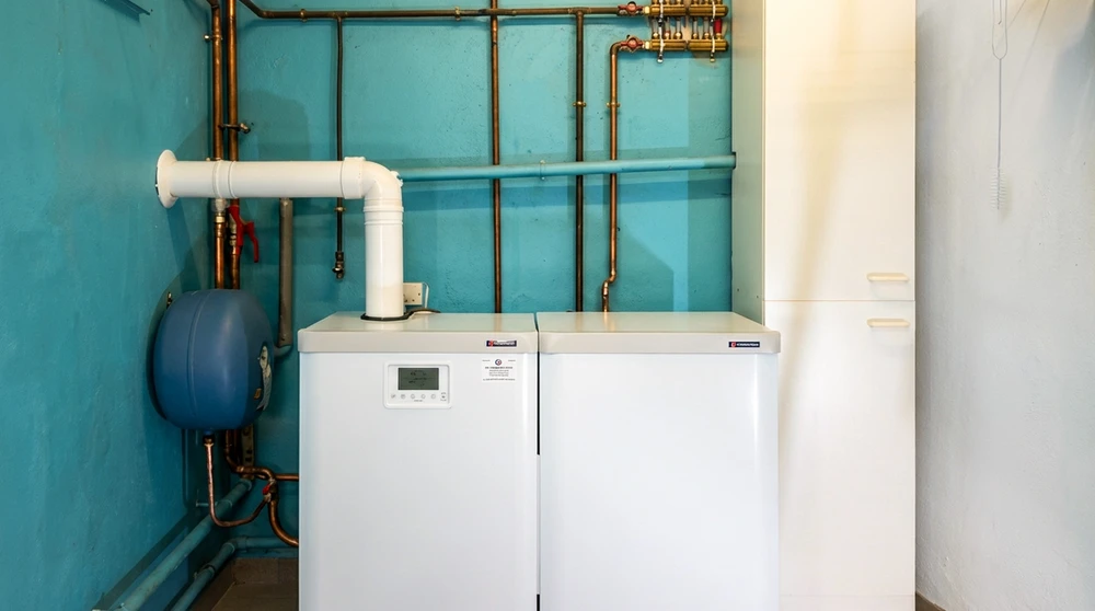 Chaufferie équipée de deux chaudières à condensation au sol blanches, marque Fröling, avec vase d'expansion bleu, collecteur de distribution en laiton, réseau de tuyauterie cuivre apparent, conduit d'évacuation PVC courbé